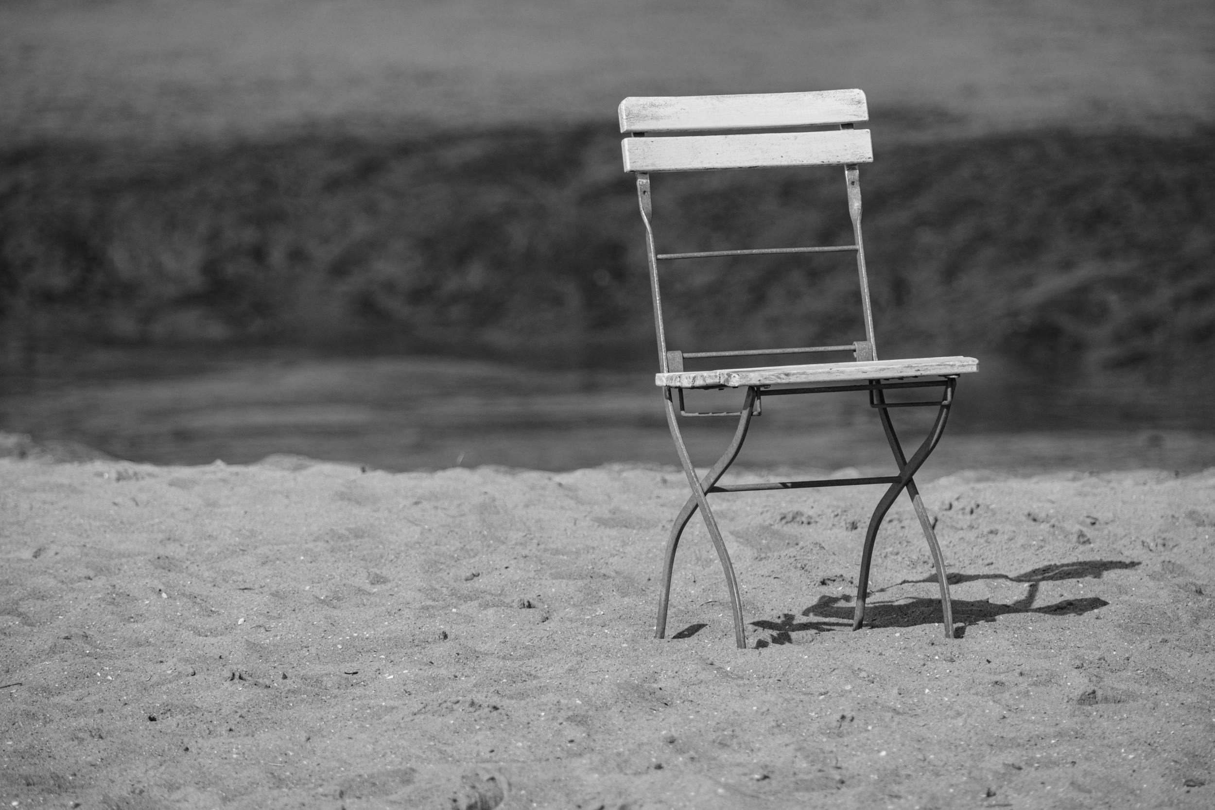 An empty wooden chair sits on a sandy beach, captured in black and white, evoking solitude.