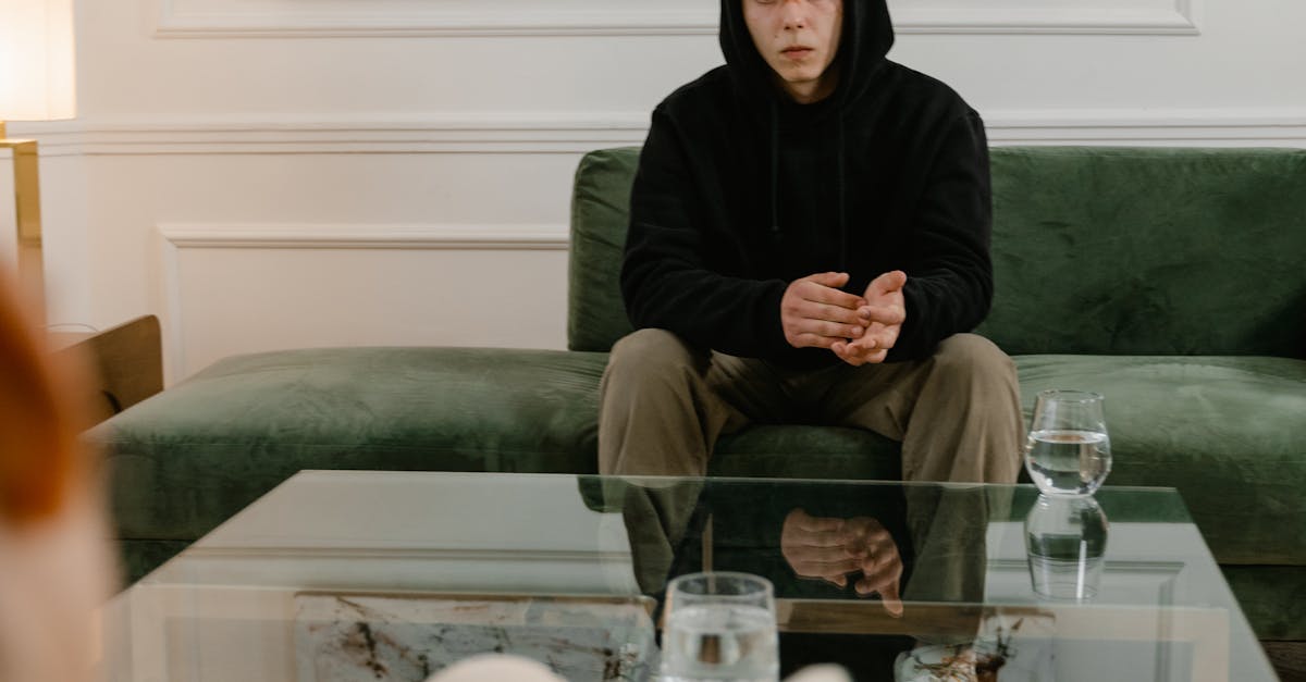 A man in a black hoodie sits on a couch, attending a therapy session indoors.